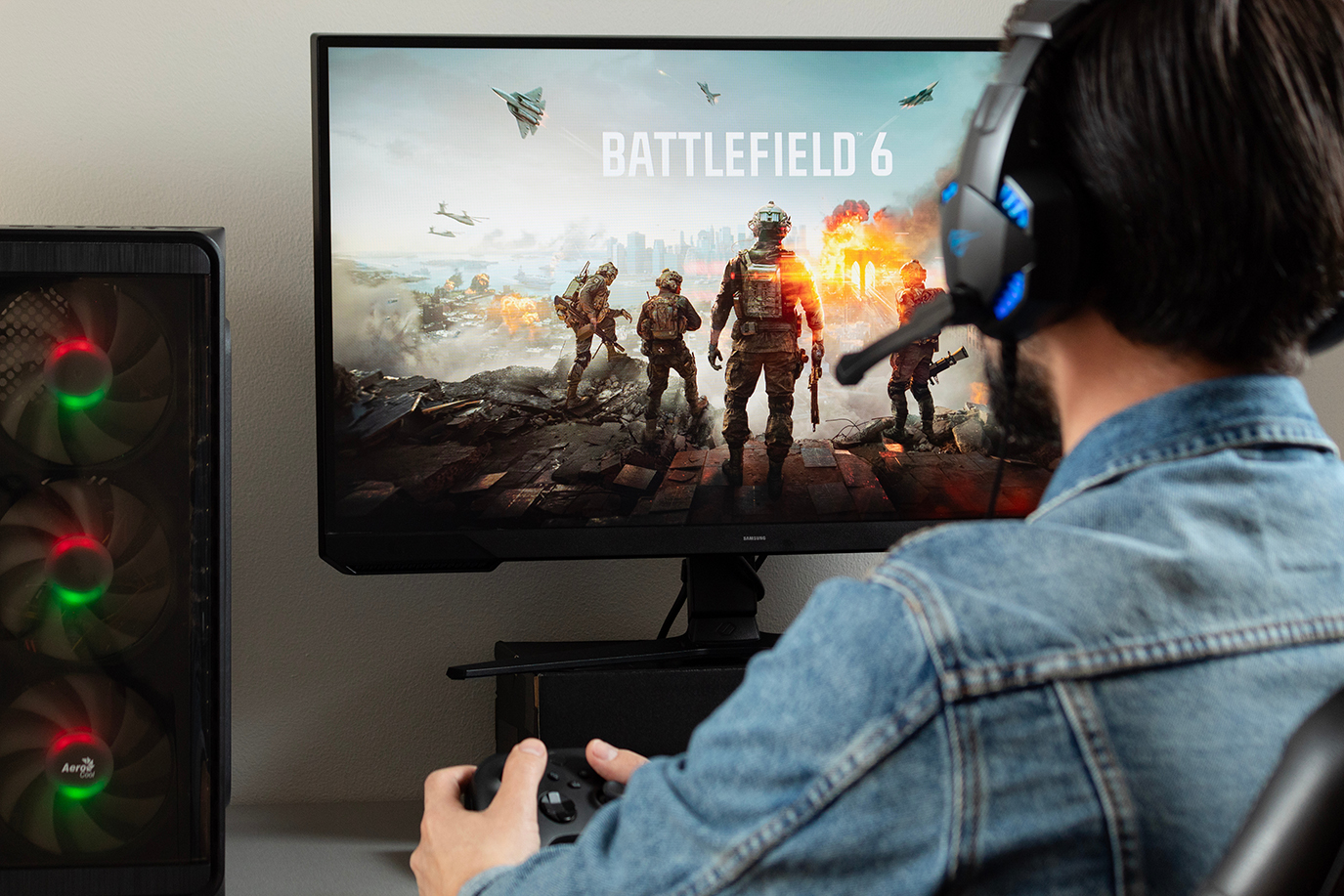 a male gamer at the right side of the picture holding a xbox controller watching a pc monitor with battlefield 6 shooter video game